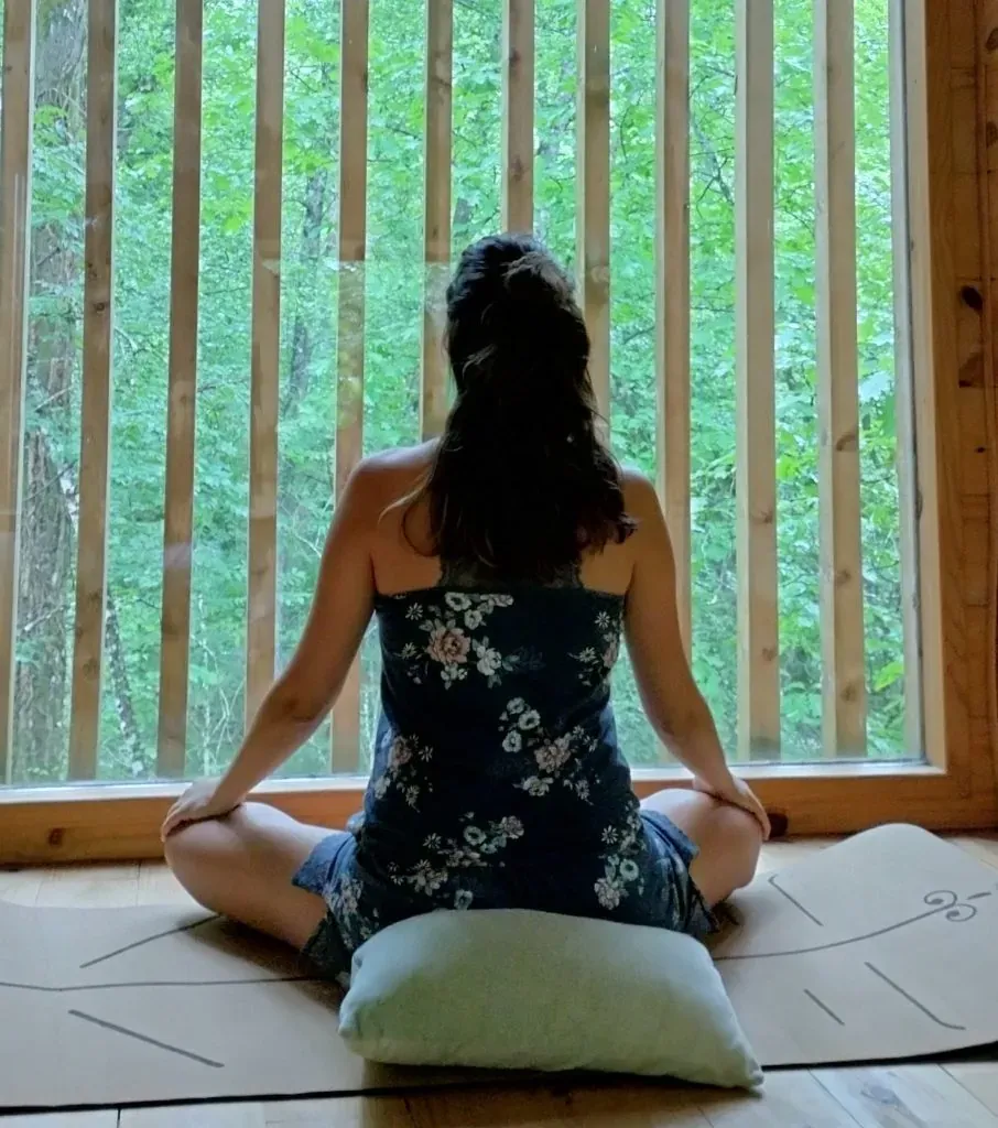 Yaiza meditando sentada sobre un cojín, frente a una ventana de listones de madera